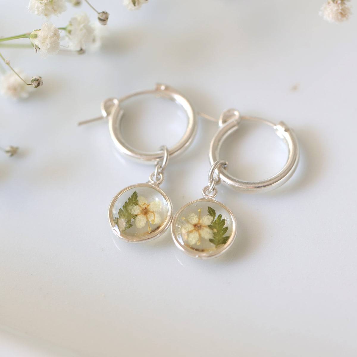 A pair of silver hoop earrings with round pendants, the pendants contain fern and black elderberry flower designs, placed on a white surface with flowers in the background.