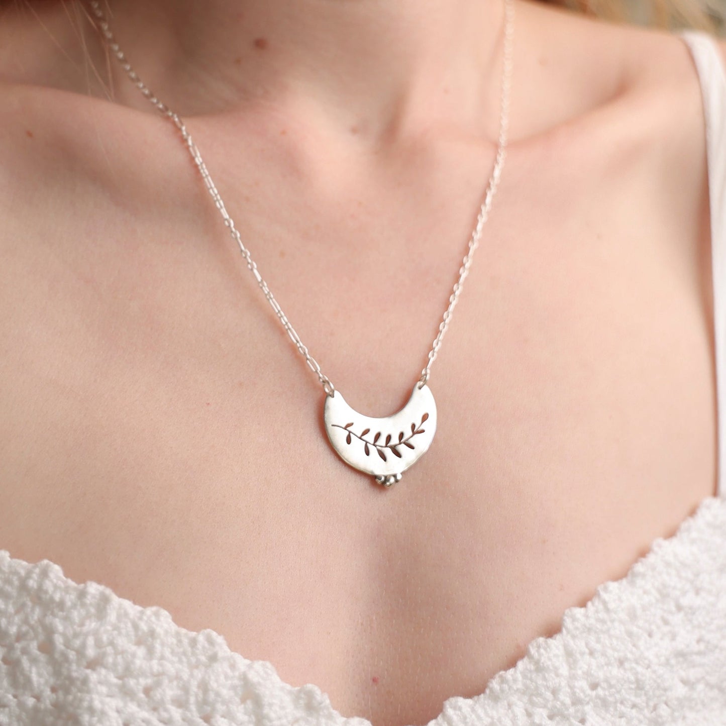 Silver necklace with a leaf pendant worn by a person in a white top, blurred natural background
