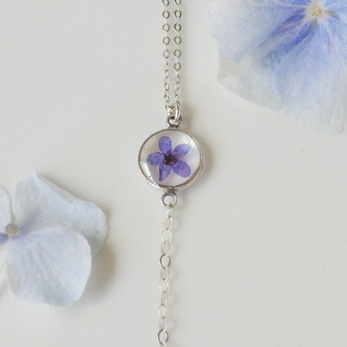 Silver necklace with a pendant featuring a pressed flower, surrounded by blue flowers on a light background