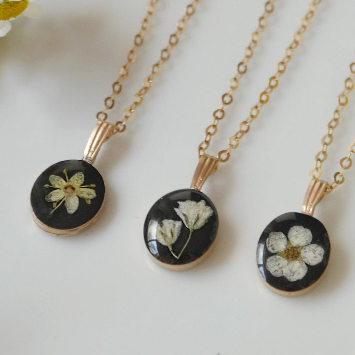 Three gold necklaces with black pendants featuring pressed flowers on a white background.