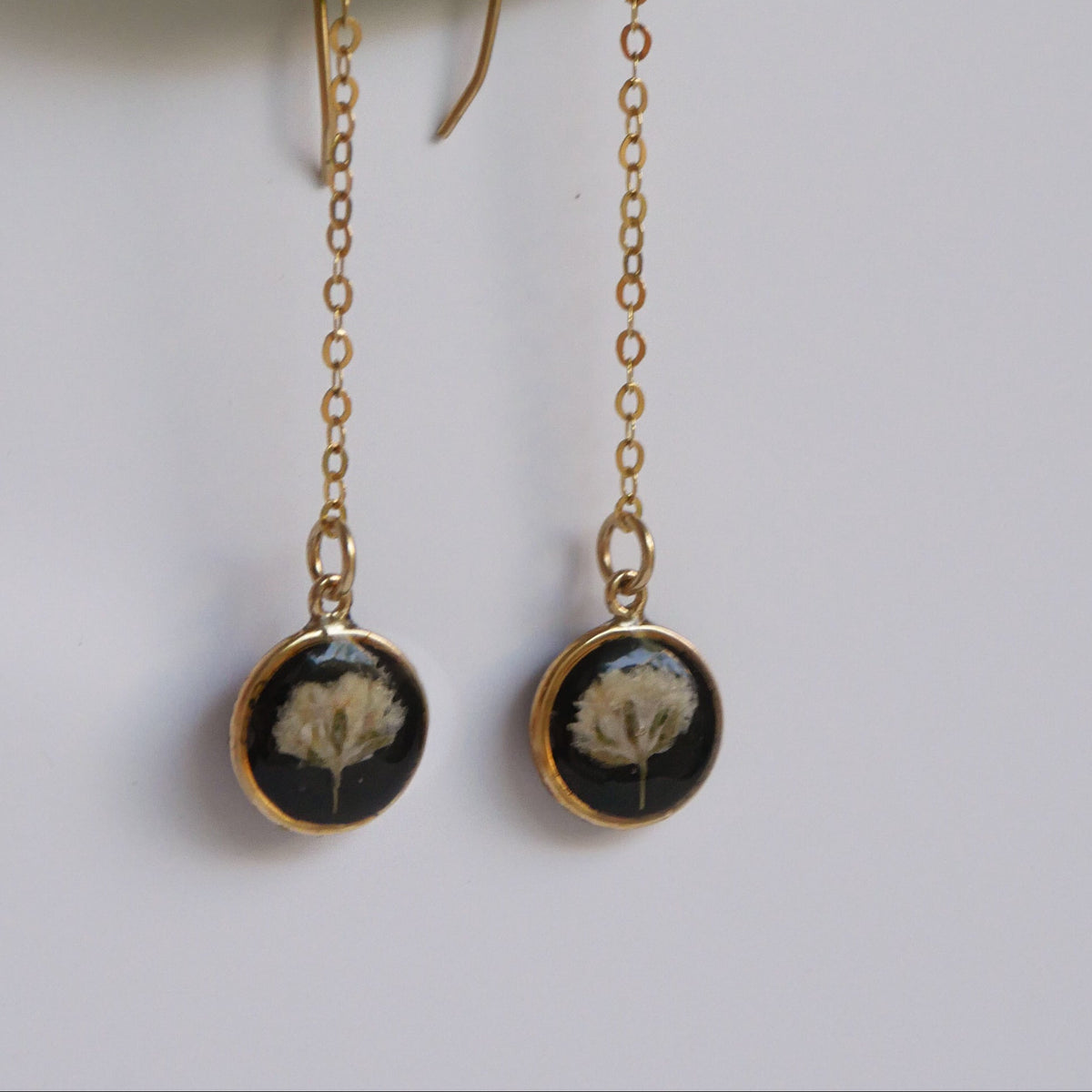 Gold chain earrings with black circular pendants featuring dried flowers, on a green leaf background.