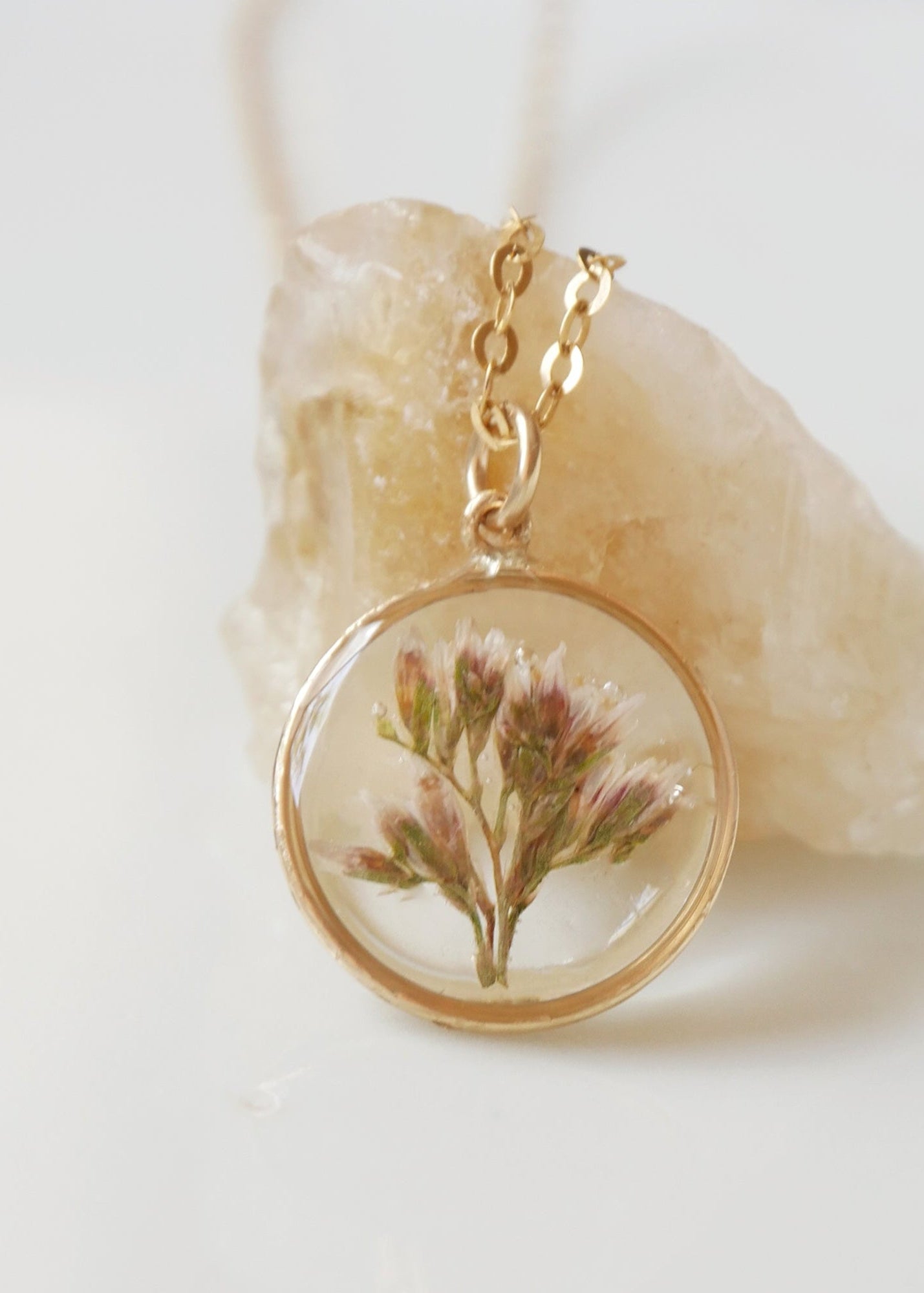Necklace with a gold chain and clear pendant featuring a pressed flower, on a light background.