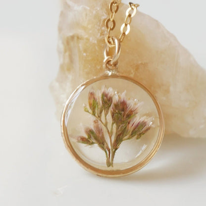 Necklace with a gold chain and clear pendant featuring a pressed flower, on a light background.