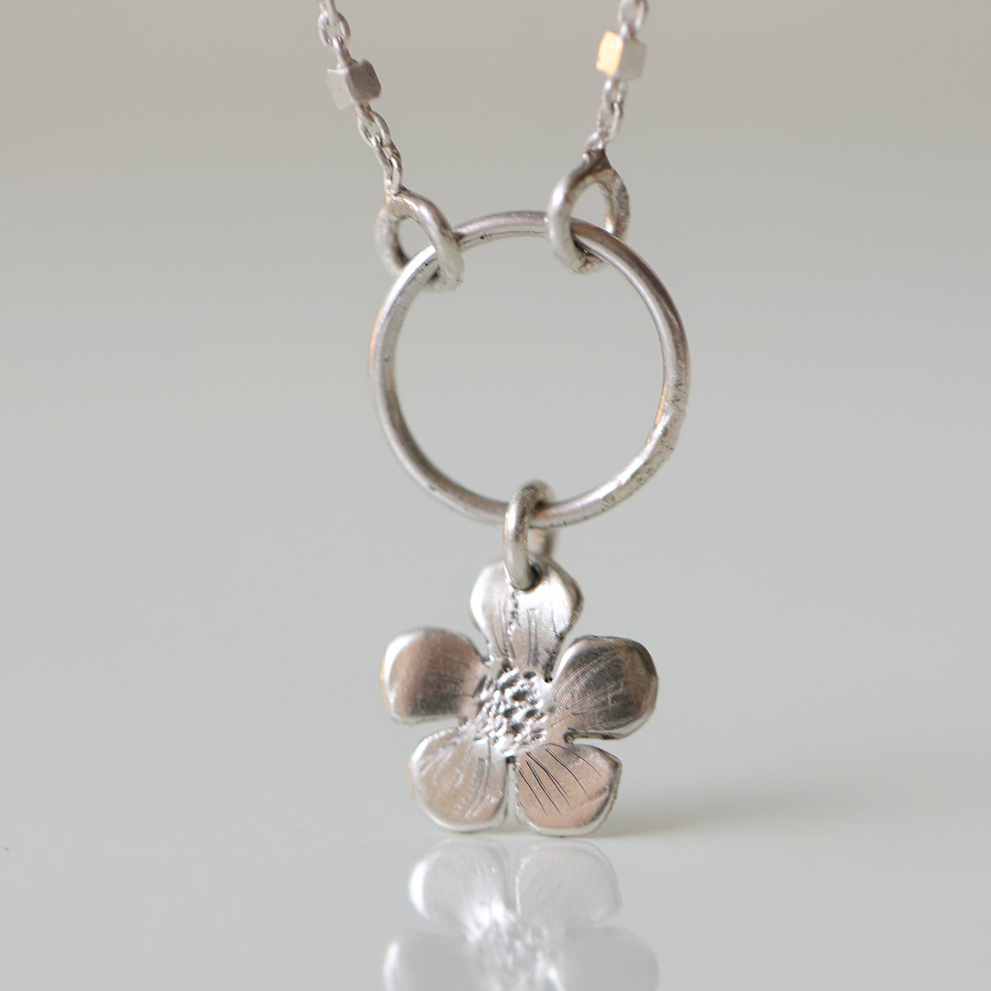 A silver necklace featuring a textured five-petal flower pendant suspended from a circular ring on a delicate chain, set against a light gray background with a soft reflection below.