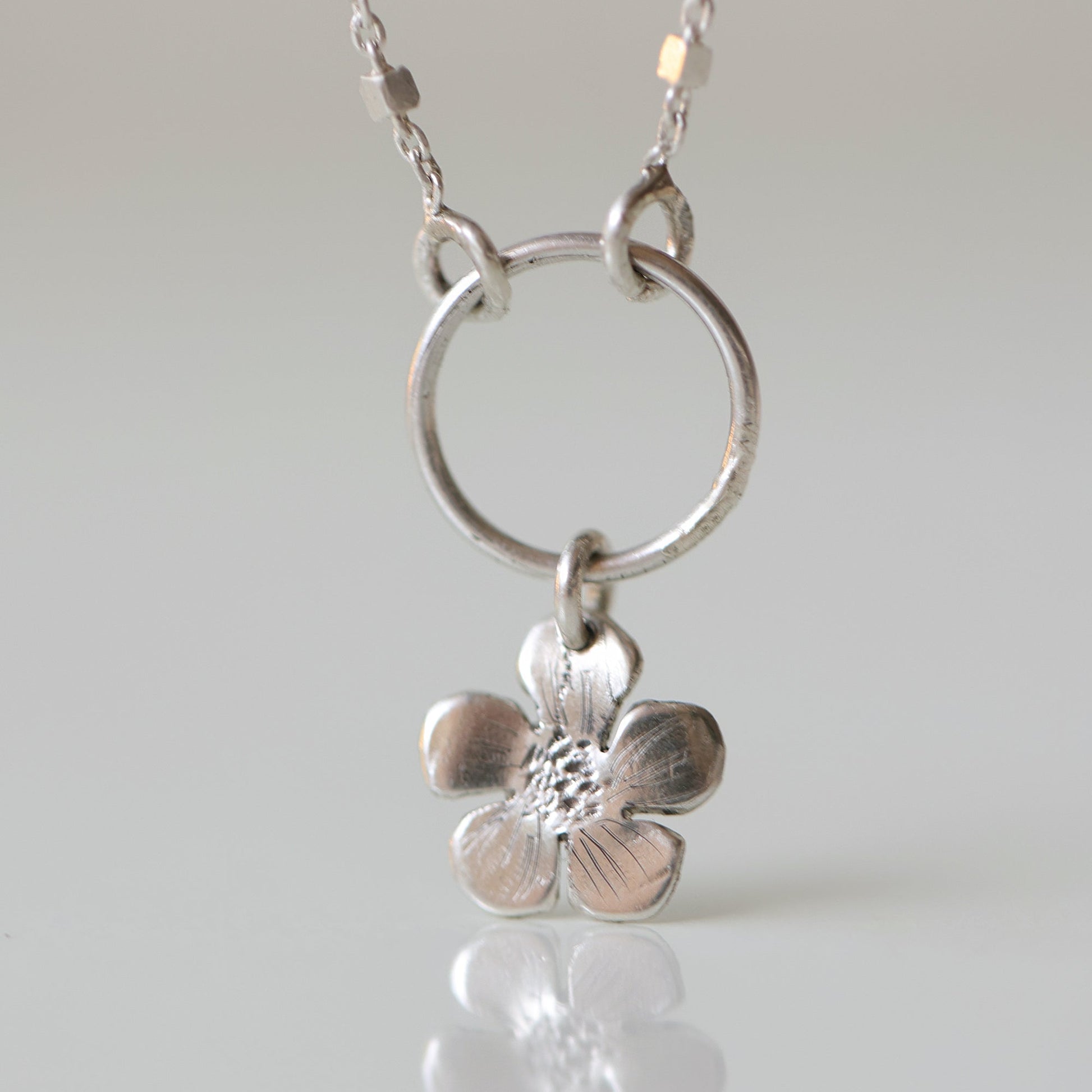 A silver necklace featuring a textured five-petal flower pendant suspended from a circular ring on a delicate chain, set against a light gray background with a soft reflection below.