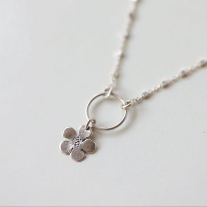A delicate silver chain necklace featuring a small flower charm with a tiny clear stone in the center, suspended from a simple silver ring, shown on a light off-white background.
