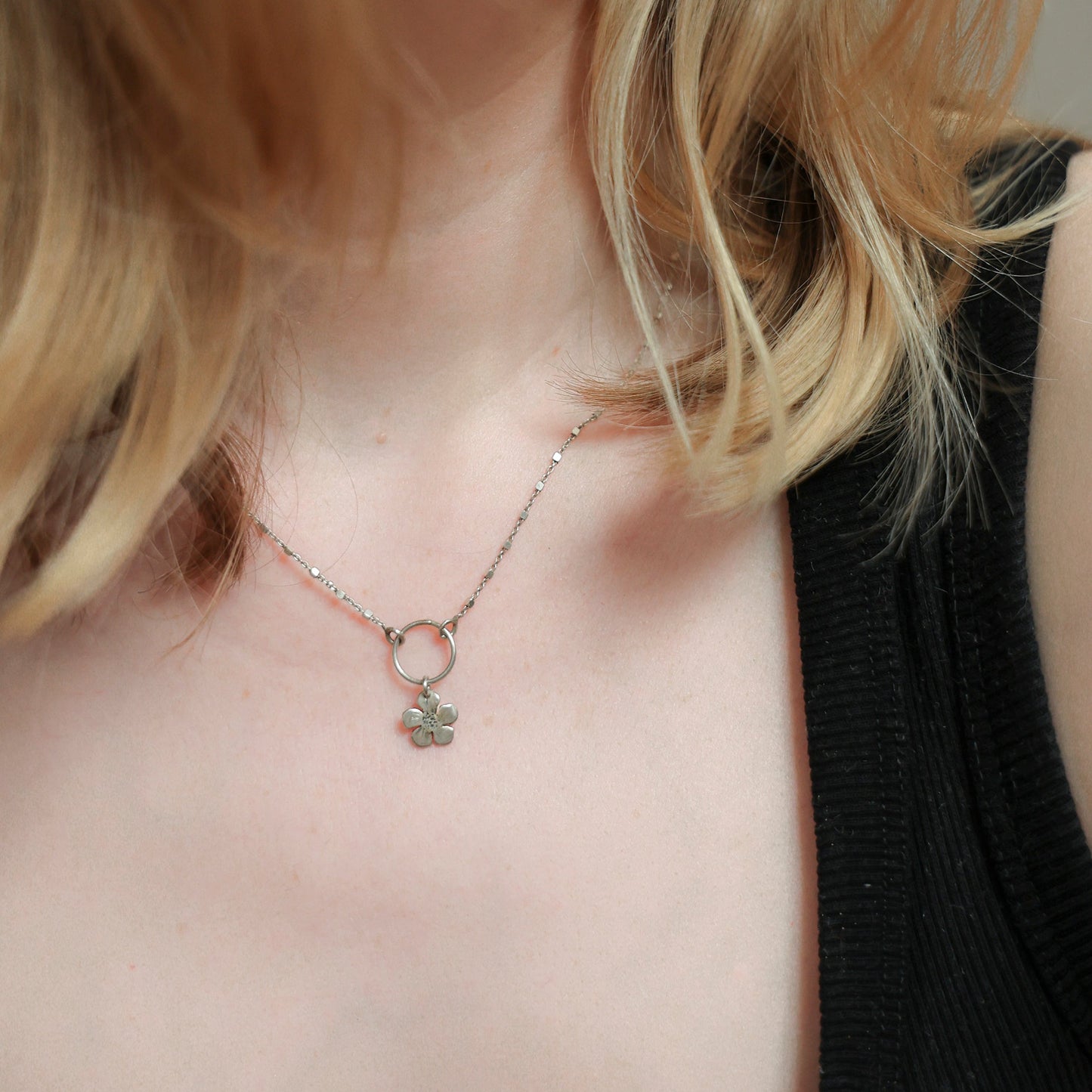 A close-up of a model wearing a delicate gold chain necklace with a small ring and clover-shaped pendant, paired with a black ribbed tank top.