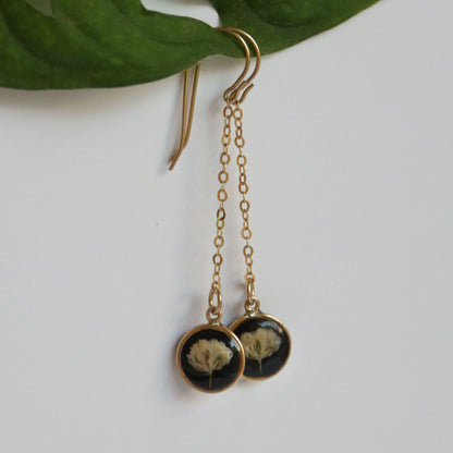 Gold chain earrings with black circular details on a white background with green leaves.