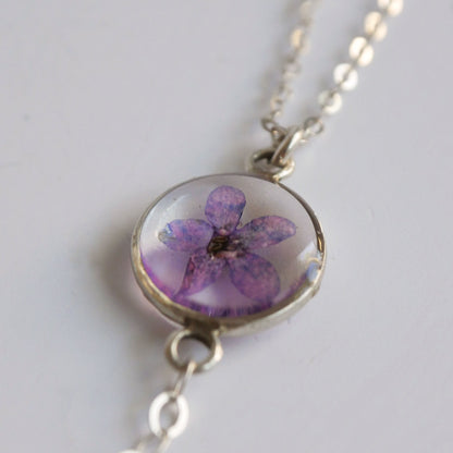 Necklace with a round pendant featuring a pressed purple flower on a light gray background
