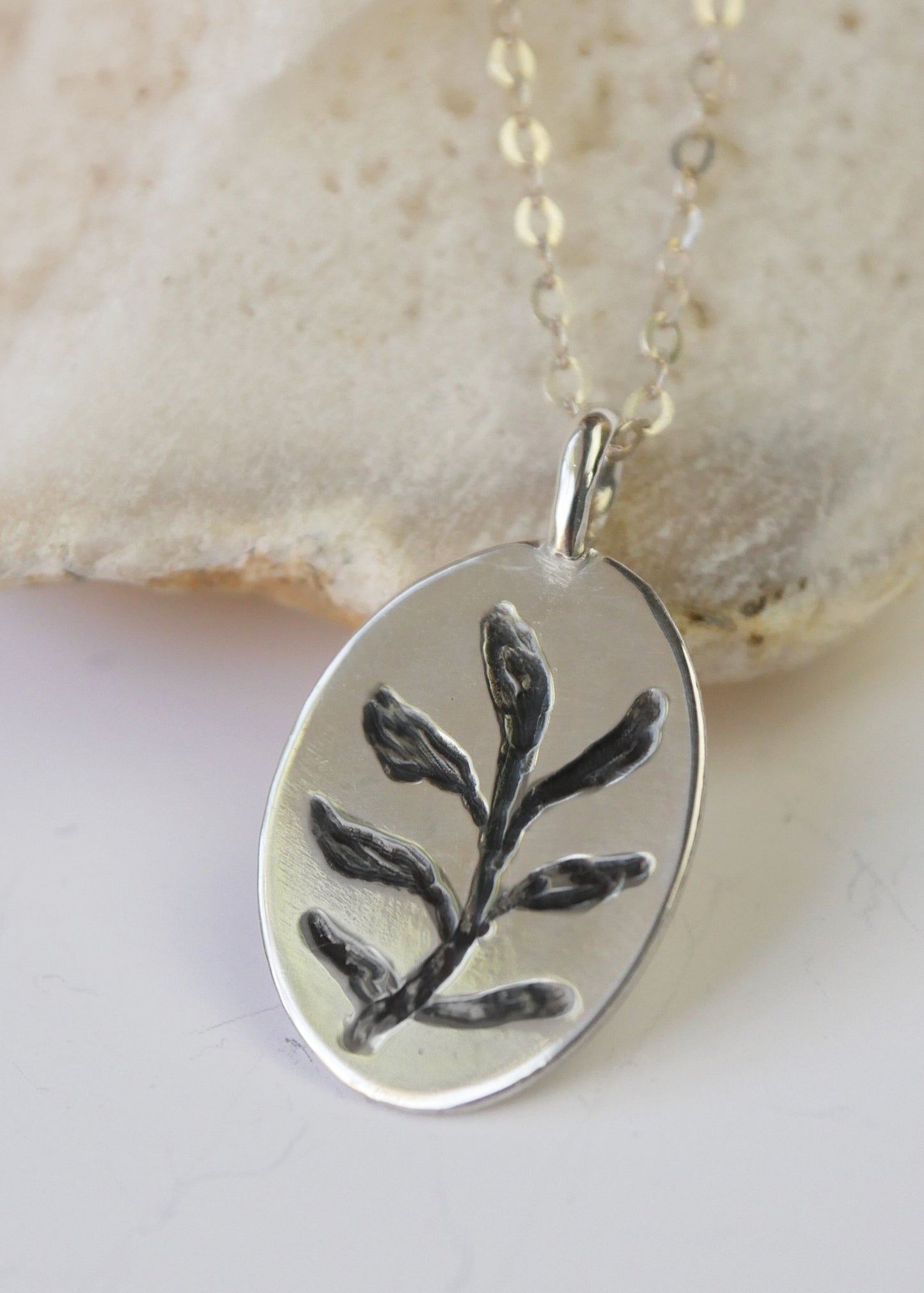 Silver pendant with leaf design on a chain, set against a beige stone background
