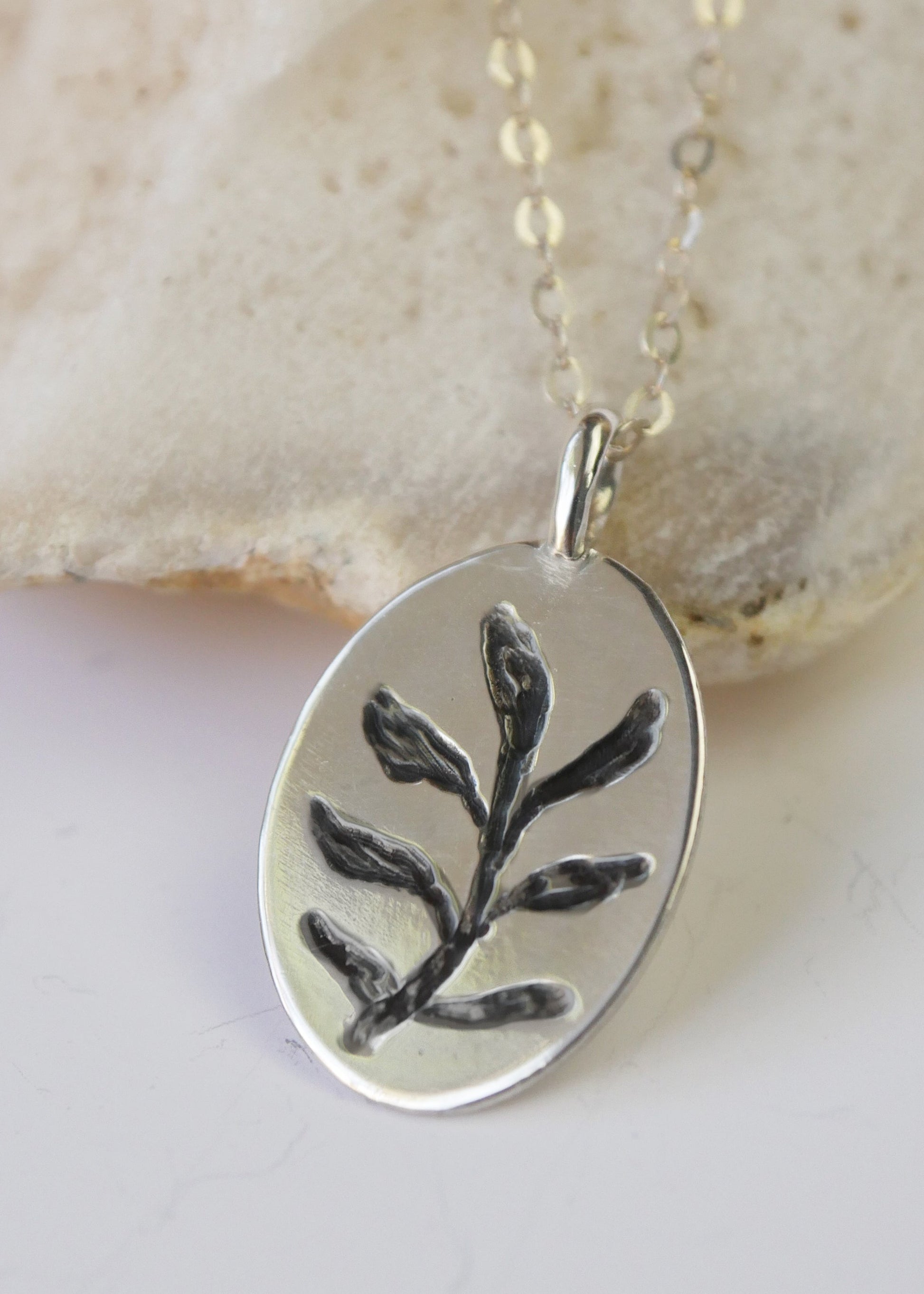 Silver pendant with leaf design on a chain, set against a beige stone background