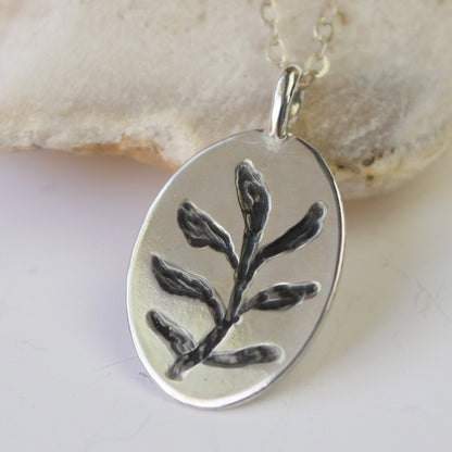 Silver pendant with leaf design on a chain, set against a beige stone background