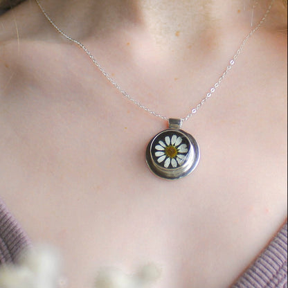 Necklace with a daisy pendant worn by a person, surrounded by white flowers.
