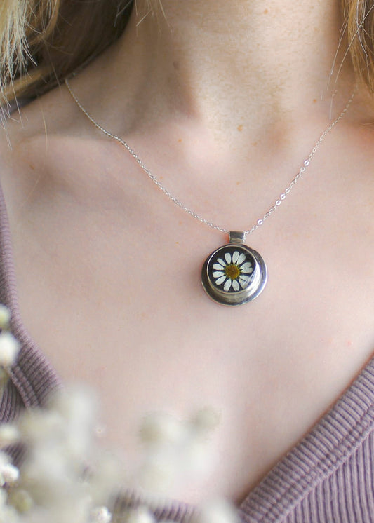 Necklace with a daisy pendant worn by a person, surrounded by white flowers.