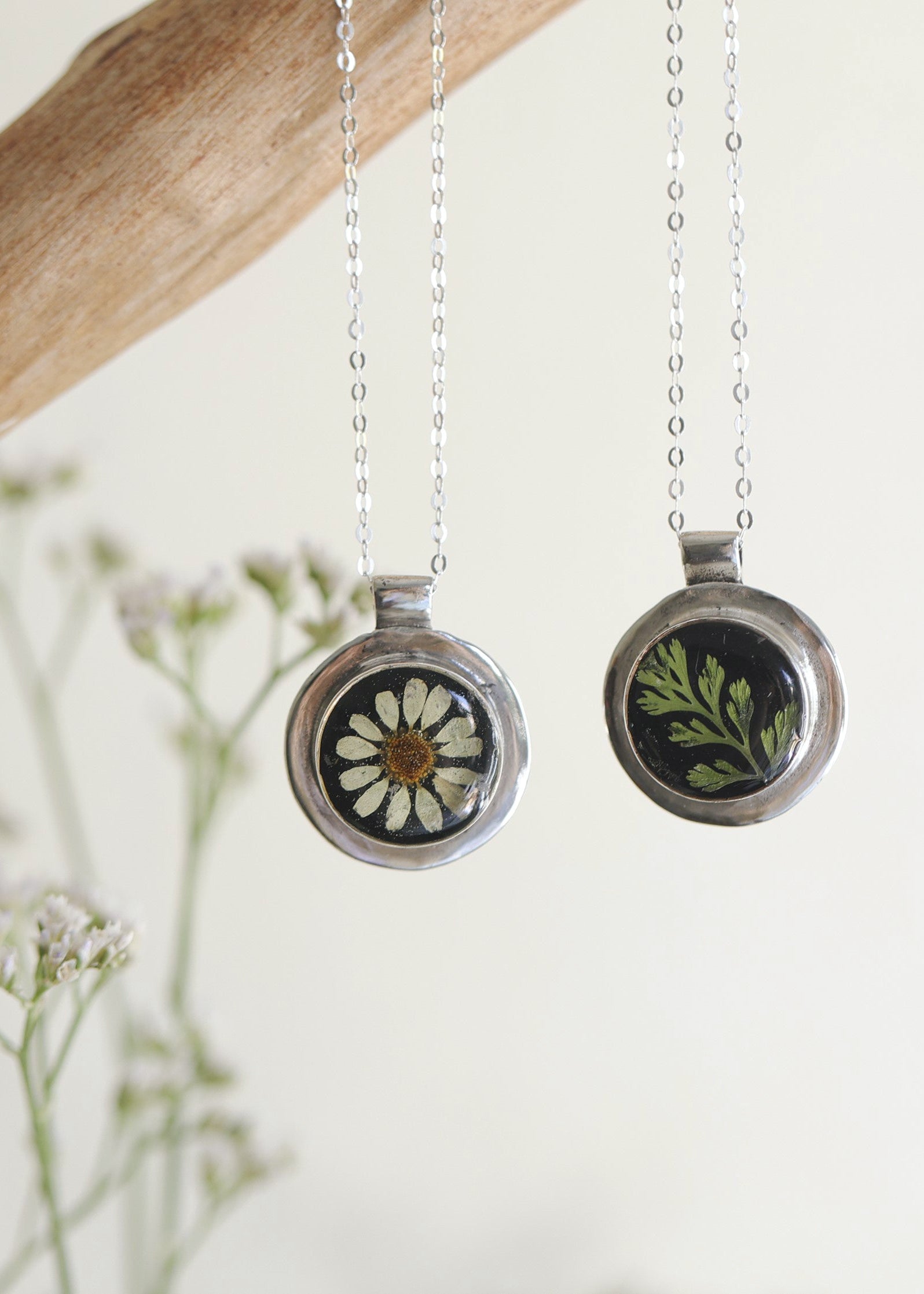 Two necklaces with round pendants, one featuring a black background with a chamomile flower, and the other with a green fern-like design, suspended by chains against a neutral background.