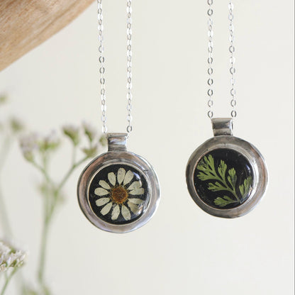 Two necklaces with round pendants, one featuring a black background with a chamomile flower, and the other with a green fern-like design, suspended by chains against a neutral background.